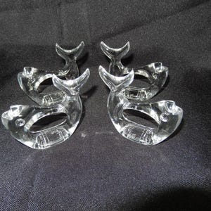 May include: Clear glass whale-shaped napkin rings. Each ring features a whale tail and a circular opening for a napkin. The rings are transparent and sit on a dark background. A set of four napkin rings.