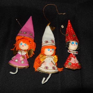 May include: Three vintage Christmas elf ornaments. Each elf has a painted wooden head, orange yarn hair, and a pointed hat. The elves wear dresses in pink, white, and red, with gold trim and dangling legs. They are suspended by gold string.