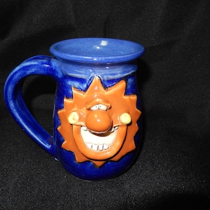 May include: A blue ceramic mug with a whimsical sun face design. The mug features a large handle and a smiling sun face in orange with white teeth. The mug is approximately 4 inches tall.
