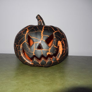 May include: A black and orange ceramic Halloween pumpkin with a cracked and weathered design. The pumpkin has a carved jack-o'-lantern face.