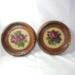 May include: Two circular wall plaques featuring needlepoint floral designs of pink roses with green leaves. Each piece is encased in a round wooden frame with a gold-toned inner rim, showcasing detailed embroidery on a cream-coloured fabric background.