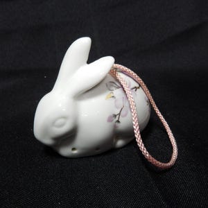 以下が含まれることがあります： ピンクの紐が付いた白いセラミック製のウサギの置物。ウサギはピンクと黄色のアクセントのフローラルデザインです。置物は黒い表面にあります。