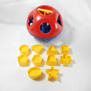 May include: A red, blue, and yellow shape sorter toy with various yellow geometric shapes. The toy is designed to help children learn about shapes and colors. The ball-shaped sorter has multiple openings.