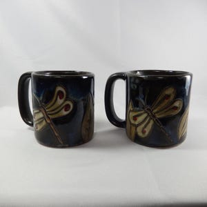 May include: Two dark blue and black ceramic mugs featuring a hand-painted dragonfly design with cream-colored wings and red accents. Each mug has a sturdy handle and a smooth, glossy finish, showcasing a nature-inspired motif on the exterior.