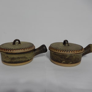 Puede incluir: Dos platos para hornear de cerámica marrón y verde con tapas y asas. Los platos tienen un aspecto texturizado y rústico.
