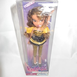 Könnte beinhalten: Eine Bratz-Puppe namens Yasmin in einer transparenten Kunststoffverpackung. Die Puppe trägt ein gelb-schwarzes Outfit im Bienen-Design, einschließlich Rock, Oberteil und gestreiften Leggings. Die Puppe hat braunes Haar und trägt silberne Schuhe. Das Bratz-Logo ist sichtbar.