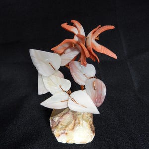May include: A handcrafted sculpture of a tree with white and pink shell leaves and coral-colored shell flowers. The tree is mounted on a beige stone base, with copper wire used to connect the leaves and flowers. The background is black.