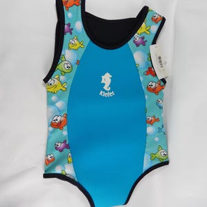Könnte beinhalten: Ein blau-türkisfarbener Baby-Badeanzug mit einem bunten Fischmuster. Der Badeanzug hat eine schwarze Einfassung und ein Seepferdchen-Logo mit dem Wort "Klefet". Der Badeanzug ist für Wasseraktivitäten konzipiert.