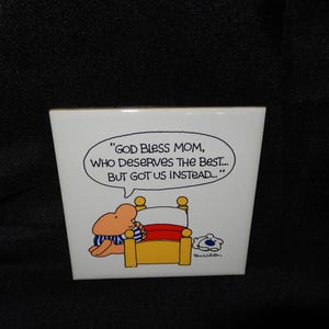 May include: A white ceramic tile with a cartoon illustration. The image features a character in blue and white striped clothing, sitting on a yellow bed with a red blanket. A speech bubble says, "GOD BLESS MOM, WHO DESERVES THE BEST... BUT GOT US INSTEAD..."