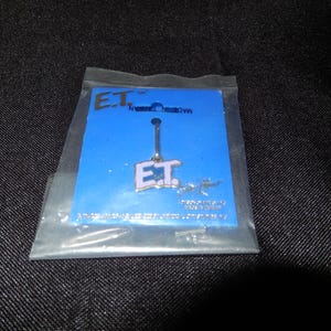May include: A silver-colored E.T. pendant with white lettering, attached to a blue card inside a clear plastic bag. The card has the text "E.T." and "MADE IN TAIWAN". The background is a dark, textured fabric.