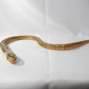 May include: A wooden snake sculpture, featuring a detailed pattern in brown and tan hues. The snake is curved, with a rounded head and a tapered tail. The sculpture is set against a white backdrop.