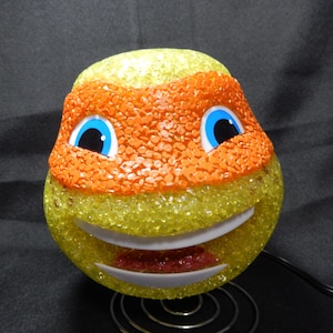 Puede incluir: Lámpara novedosa con forma de cabeza de personaje de dibujos animados, con una combinación de colores amarillo y naranja. El personaje tiene ojos azules, boca blanca y lengua roja. La lámpara está sobre un soporte metálico.