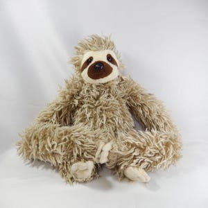 May include: A plush sloth toy featuring long, shaggy light brown fur, a cream-coloured face with dark brown eye patches, and a dark brown nose. The stuffed animal has long arms and legs with light-coloured paws, sitting in a relaxed position.