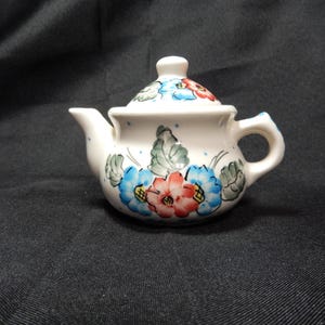 Op de afbeelding: Kleine, witte keramische theepot met deksel. De theepot is versierd met blauwe, rode en groene bloemmotieven. Hij heeft een klein handvat en een tuit. De theepot wordt waarschijnlijk gebruikt voor het serveren van thee.