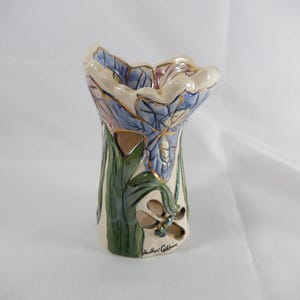 May include: A ceramic vase shaped like a flower, with blue, pink, and gold accents. The vase has green leaves and a dragonfly cutout. The artist's signature, "Heather Goldminc," is visible on the base. The vase is a decorative item.