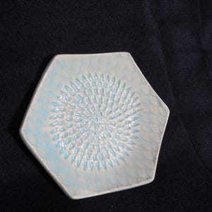 Puede incluir: Un rallador hexagonal de cerámica en azul claro y blanco. El centro presenta un diseño en espiral texturizado, rodeado por un borde azul claro. Este utensilio de cocina está diseñado para rallar ingredientes.