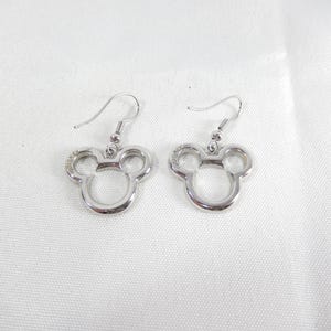 K&ouml;nnte beinhalten: Ein Paar silberfarbene Ohrh&auml;nger mit einer ausgeschnittenen Silhouette eines Mauskopfes mit abgerundeten Ohren. Jeder Ohrring ist an einem gebogenen Metallhaken befestigt und die Metalloberfl&auml;che weist ein glattes, reflektierendes Finish auf.