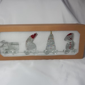 May include: A clear glass Christmas train set featuring a locomotive, snowman, Christmas tree, and another snowman, all with red and colourful accents. The set is presented in a brown cardboard box with a white foam insert.