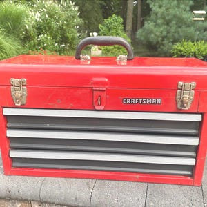 May include: A red metal Craftsman tool box featuring a black handle, two silver-toned metal latches, and three horizontal drawers with silver-toned trim. The brand name Craftsman is printed in white text on a black rectangular background.