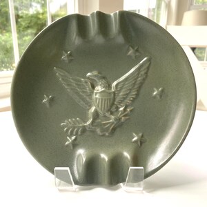 May include: A round, sage green ceramic ashtray featuring a central embossed eagle emblem with a shield on its chest, clutching arrows. The design includes four small embossed stars surrounding the eagle and three integrated cigarette rests.