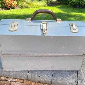 May include: A rectangular gray metal tool box featuring a dark brown handle on top and three silver-colored metal latches across the front lid. The exterior surface shows signs of wear with minor scratches and small orange spots of oxidation.