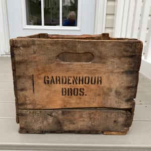 May include: A weathered wooden crate with the text "GARDENHOUR BROS." in black stencil lettering. The crate has a rectangular shape with a handle cut out on one side. The wood has a natural, aged appearance.