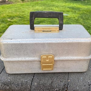 May include: A silver-colored, rectangular toolbox with a black handle and a gold-colored clasp. The toolbox has a textured surface and a hinged lid. The handle is attached to the top of the box with gold-colored hardware.