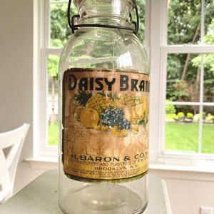 May include: A clear glass jar with a metal handle and lid. The jar has a label with the text "DAISY BRAN" and illustrations of fruits. The label also includes the text "H. BARON & CO. BROOKLYN, N.Y."