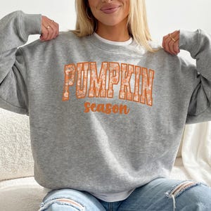 Peut inclure: Un sweat-shirt gris chiné à col rond avec le texte PUMPKIN en grande police universitaire orange, rempli d'un motif floral blanc, et le mot season écrit en dessous dans une police cursive orange plus petite et unie.