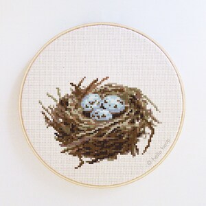 May include: A cross-stitch embroidery of a bird's nest with blue eggs. The nest is made of twigs and is surrounded by green leaves. The embroidery is framed in a round wooden hoop. The text "© hello hoop" is in the lower right corner of the embroidery.