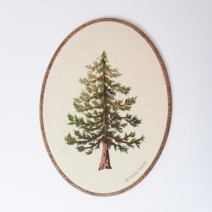 以下が含まれることがあります： 白いキャンバスに緑色の松の木が刺繍されています。茶色の木製の輪が付いており、右下に「hello hoop」の文字があります。