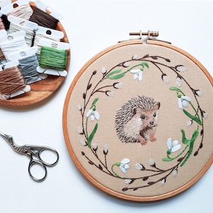 Könnte beinhalten: Stickerei-Bild in einem Reifen mit einem braunen Igel mit weißen Blüten und grünen Blättern auf beigefarbenem Hintergrund.