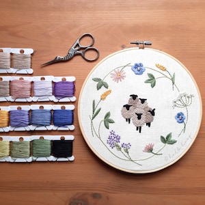 May include: A close-up of a white embroidery hoop with a floral design and a group of sheep embroidered in the centre. The hoop is surrounded by a variety of colourful embroidery floss spools.