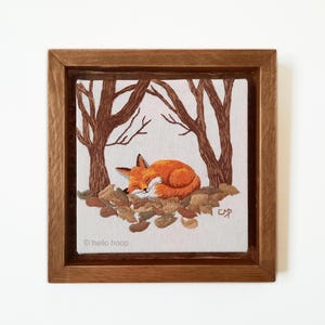 May include: Embroidered artwork of a fox sleeping in a bed of autumn leaves under two trees. The artwork is framed in a brown wood frame. The text "hello hoop" is in the bottom left corner of the artwork.