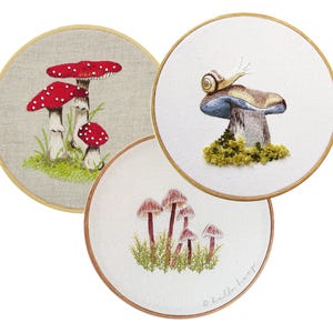 May include: Three embroidered mushroom art pieces in wooden hoops. One features red and white spotted mushrooms on a linen background. Another shows a snail on a mushroom. The third depicts pink mushrooms. The embroidery is detailed and colorful.
