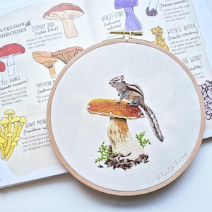 Puede incluir: Un bordado en bastidor con una ardilla listada sobre una seta. La seta tiene un sombrero marrón y un tallo beige, con hojas verdes. La obra de arte está sobre un fondo de tela blanca.