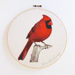 May include: A cross-stitch embroidery of a vibrant red cardinal bird perched on a branch. The bird has a black face and is set against a white background within a wooden embroidery hoop. The artist's signature is visible.