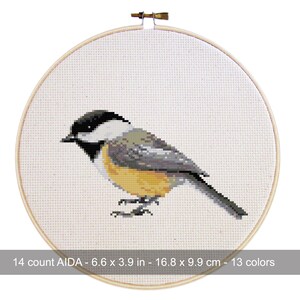 May include: A cross-stitch embroidery of a chickadee bird, featuring black, white, gray, and yellow hues. The bird is centered on a white AIDA cloth, framed by a wooden hoop. The text at the bottom reads: 14 count AIDA - 6.6 x 3.9 in - 16.8 x 9.9 cm - 13 colors.