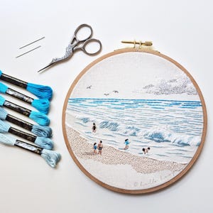 Puede incluir: Una escena de playa bordada en un aro de madera. La obra de arte representa una playa con gente en el agua, olas y un cielo nublado. Hilos de bordar en tonos azules y unas tijeras están junto al aro.