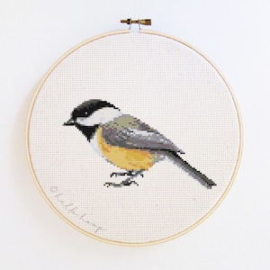 May include: A cross-stitch embroidery of a chickadee bird in a wooden hoop. The bird has a black head, white cheeks, gray back, and yellow breast. The text "hello hoop" is written in cursive on the bottom left of the fabric.