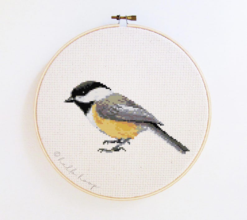 May include: A cross-stitch embroidery of a chickadee bird in a wooden hoop. The bird has a black head, white cheeks, gray back, and yellow breast. The text "hello hoop" is written in cursive on the bottom left of the fabric.