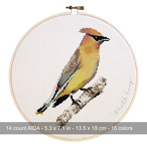 May include: A cross-stitch embroidery of a bird with yellow, brown, and black plumage perched on a branch. The artwork is framed in a wooden hoop. The text on the image reads: 14 count AIDA - 5.3 x 7.1 in - 13.5 x 18 cm - 15 colors.