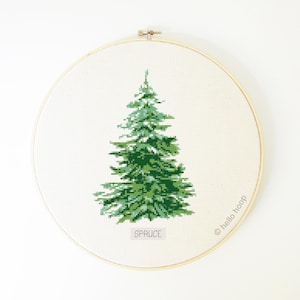 Peut inclure: Un motif de point de croix représentant un épicéa vert dans un cercle de broderie en bois. Le motif est intitulé "SPRUCE" et le cercle porte le texte "@hello_hoop" sur le côté.