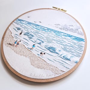 May include: Embroidered beach scene in a wooden hoop. The artwork depicts a sandy shore, ocean waves, and figures in the water. The color palette includes shades of blue, beige, and white. The text "hello hoop" is at the bottom.