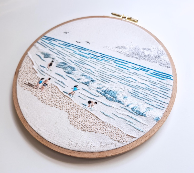 May include: Embroidered beach scene in a wooden hoop. The artwork depicts a sandy shore, ocean waves, and figures in the water. The color palette includes shades of blue, beige, and white. The text "hello hoop" is at the bottom.