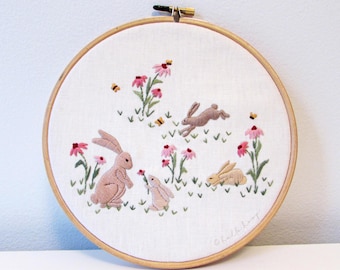 May include: Embroidered hoop art featuring three brown rabbits in a field of pink flowers. The rabbits are surrounded by green leaves and small white flowers. The hoop is made of wood and has a white background.