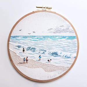 May include: Embroidered beach scene in a wooden hoop. The artwork depicts a beach with people in the water, waves, and seagulls. The sky is light blue with a few clouds. The sand is tan and the water is shades of blue.