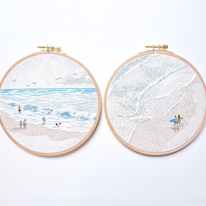 May include: Two embroidered beach scenes in wooden hoops. One shows people playing in the ocean waves, the other depicts a surfer with a surfboard. The artwork features blue, white, and tan colors, with the artist's signature at the bottom.