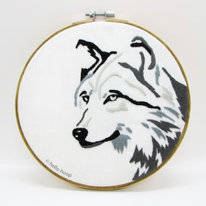 May include: Embroidered hoop art featuring a black and white wolf head with gray fur details. The hoop is made of wood and has a white fabric background. The text "hello hoop" is printed on the bottom of the hoop.