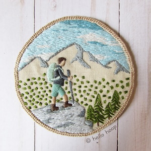 May include: Embroidered patch featuring a hiker with a backpack and walking stick, standing on a mountaintop with a view of a mountain range and a blue sky with clouds. The patch is surrounded by a brown border.